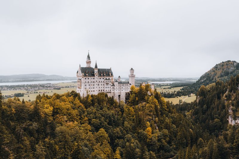 Castelo Neuschwanstein em pedra branca sobre colina coberta de floresta no outono com lago e planície ao fundo na Bavária, Alemanha