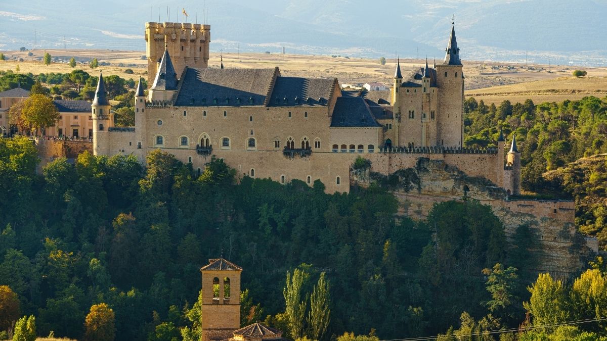Vista frontal e imponente do Alcázar de Segóvia, um castelo medieval de pedra clara na Espanha, com suas icônicas torres circulares de telhados cônicos e pontiagudos escuros sob um céu azul claro.