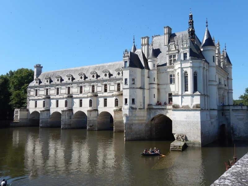 Vista lateral do Castelo de Chenonceau, na França, construído como uma ponte sobre as águas calmas do rio Cher, que refletem a fachada renascentista branca da estrutura sob a luz do entardecer.