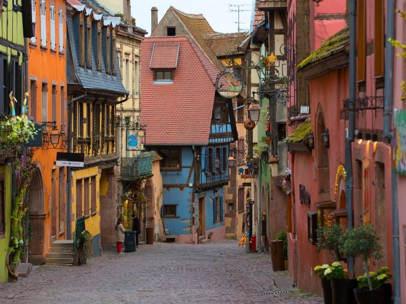 Uma rua de paralelepípedos estreita na vila medieval de Riquewihr, na França, ladeada por casas coloridas em estilo enxaimel com floreiras nas janelas e uma torre de pedra ao fundo.
