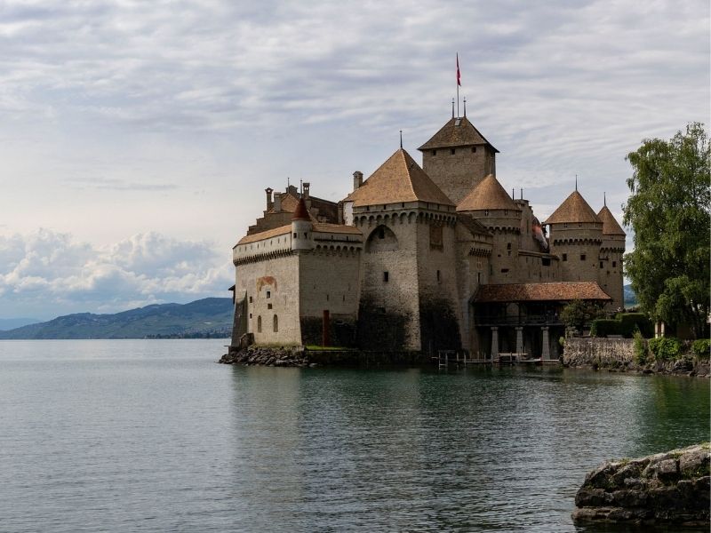 O medieval Castelo de Chillon situado às margens rochosas do azul e límpido Lago Leman, na Suíça, com montanhas imponentes e áreas arborizadas ao fundo sob um céu com nuvens suaves.