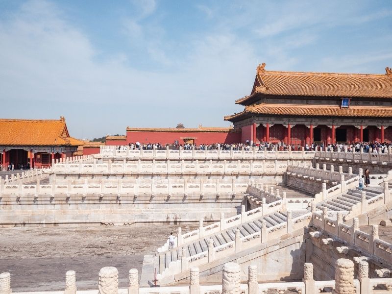 Vista frontal do Salão da Suprema Harmonia na Cidade Proibida, exibindo a arquitetura tradicional chinesa com telhados duplos em tons de amarelo e vermelho, sob um céu azul e limpo.