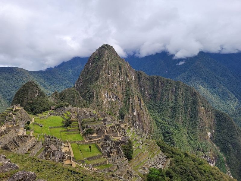 Vista panorâmica e elevada das ruínas de pedra da antiga cidadela inca de Machu Picchu, no Peru. O complexo arqueológico está situado entre montanhas verdes e imponentes, com o pico do Huayna Picchu ao fundo sob um céu com nuvens esparsas.