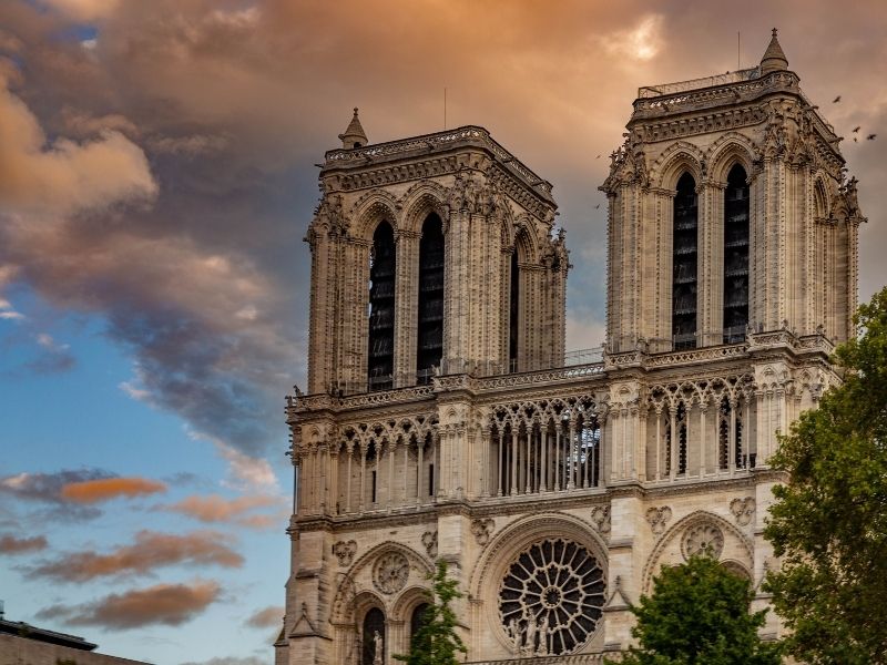Fachada posterior da Catedral de Notre-Dame em Paris vista a partir do rio Sena durante o pôr do sol, com tons dourados e rosados no céu iluminando a arquitetura gótica e as torres.