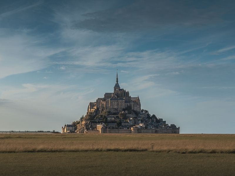 Vista frontal da Abadia do Mont Saint-Michel, com sua imponente torre central e detalhes de arquitetura gótica e românica, destacando-se no topo da ilha rochosa sob um céu nublado.