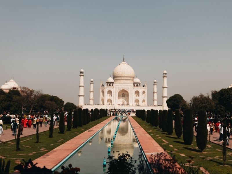 Vista frontal do Taj Mahal sob um céu azul claro. O imponente mausoléu de mármore branco exibe sua grande cúpula central, arcos simétricos e minaretes nas laterais, com o jardim e o espelho d'água em primeiro plano.