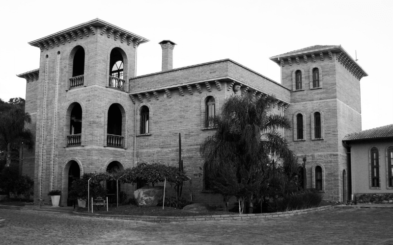 Fachada em preto e branco do Castelo Casa Valduga, construção de pedra com torres e arcos, rodeada de vegetação e pátio de paralelepípedos.