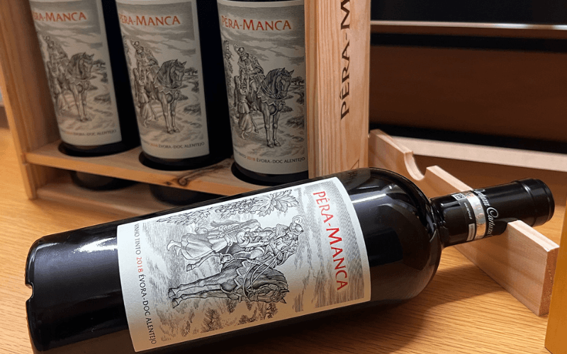 Garrafa do vinho Pera-Manca tinto 2018 deitada sobre caixote de madeira com outras garrafas da mesma etiqueta ao fundo.