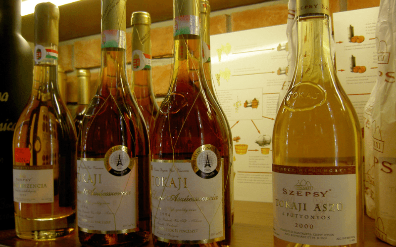 Garrafas de vinho Tokaji húngaro enfileiradas, incluindo rótulos Tokaji Aszú e Szepsy, com painel explicativo ao fundo.