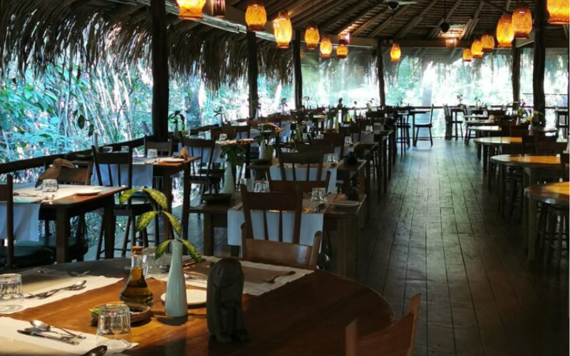 Restaurante do Anavilhanas Jungle Lodge com telhado de palha, mesas de madeira, luminárias de vime alaranjadas e vegetação amazônica ao redor.
