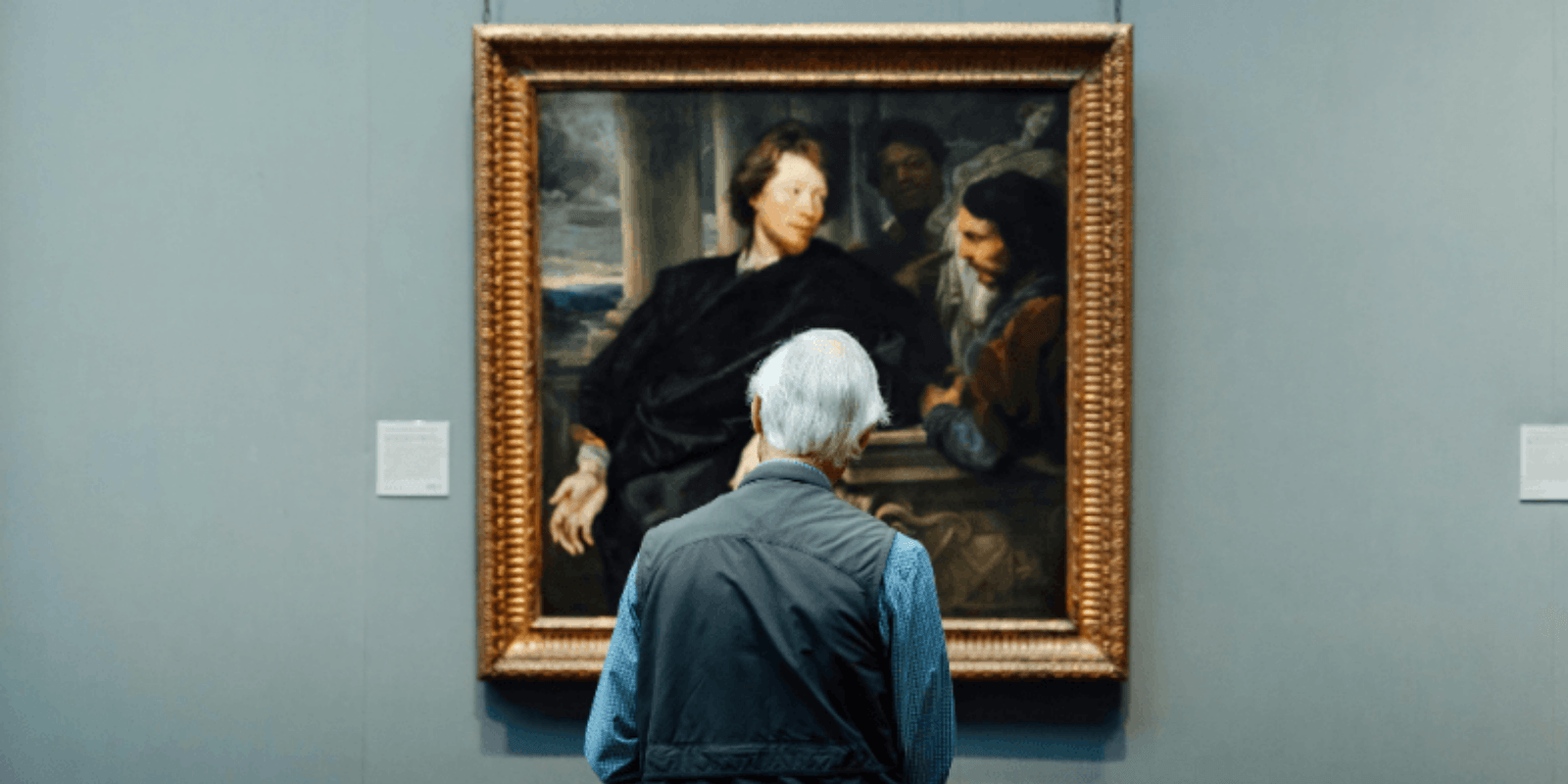Homem idoso de costas observando uma pintura clássica em moldura dourada em um museu.