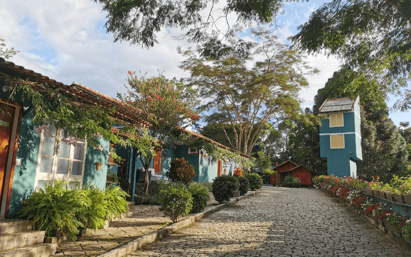 Alameda de paralelepípedos da Pousada Rabo do Lagarto com casarios azuis, jardins floridos e árvores frondosas em Pedra Azul, ES.