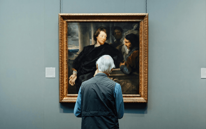 Homem idoso de costas observando uma pintura clássica em moldura dourada em um museu.