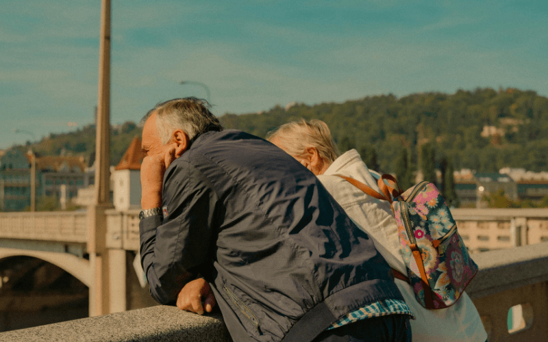 Casal sênior apoiado em um muro observando a paisagem urbana com ponte e colina ao fundo.