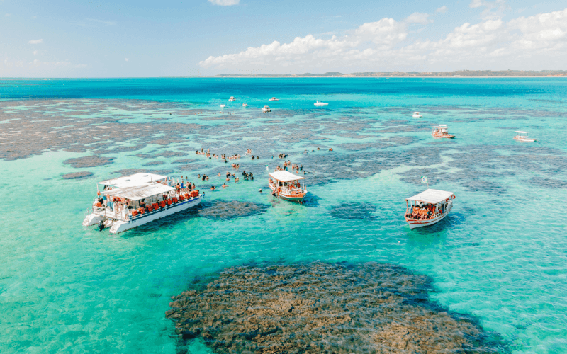 Vista aérea de barcos turísticos e pessoas nas piscinas naturais de mar turquesa com recifes de coral visíveis.