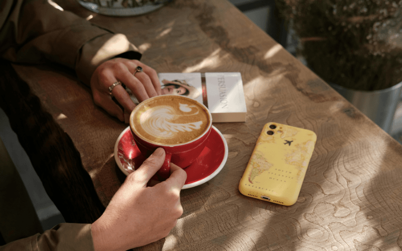 Pessoa segurando xícara vermelha de café com latte art em mesa de madeira ao lado de celular com capinha de mapa-múndi.