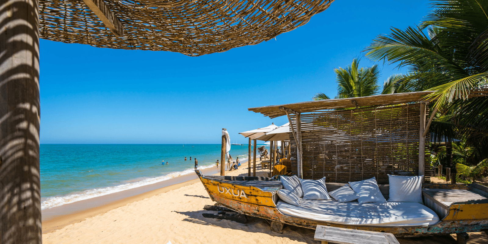 Área de descanso do hotel Uxua em Trancoso com barco de madeira convertido em sofá na areia, palmeiras e mar azul ao fundo.