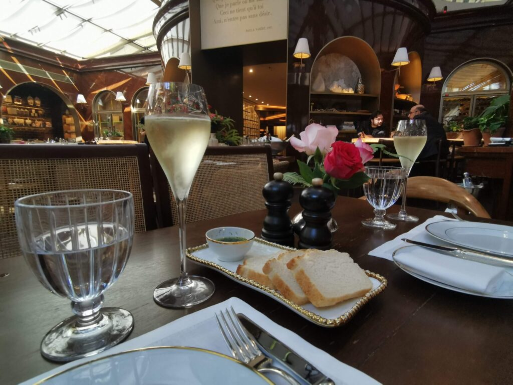 Mesa posta em restaurante elegante com taças de espumante, pão fatiado, flores rosas e decoração clássica ao fundo.
