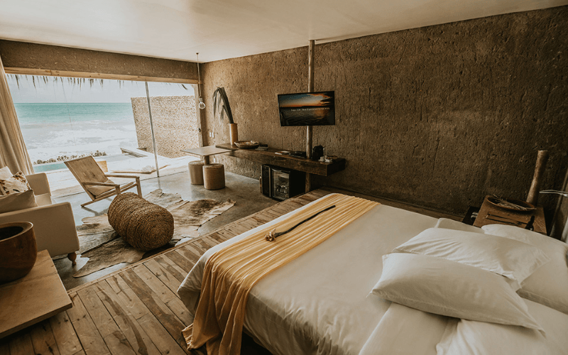 Suíte do resort Kenoa com cama de casal, paredes de concreto aparente e varanda com vista direta para o mar turquesa.