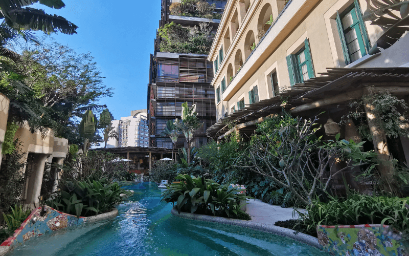 Área externa do Rosewood São Paulo com piscina sinuosa, jardins exuberantes e torre com jardins verticais em cada andar ao fundo.