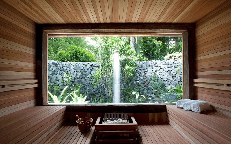 Interior de sauna em madeira com grande janela para jardim com chuveiro externo, toalhas enroladas e balde de sauna.